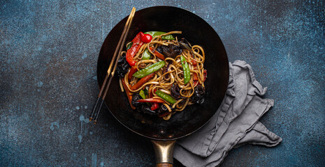 Asian dish stir fry udon noodles with vegetables and mushrooms in black rustic wok pan with wooden chopsticks on rustic dark blue concrete background from above, Chinese fast food