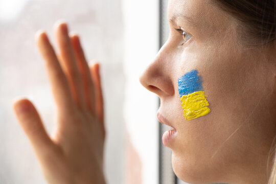 A Ukrainian woman with the national flag of Ukraine yellow-blue on her face looks out the window, patriot, peace in Ukraine, martial law, I see the war outside the window, stop war