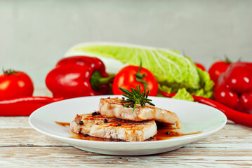 Pieces of meat grilled. Steak with rosemary and pepper in a white plate. Meat dish with red sauce on a white wooden table. Fresh tomatoes, peppers and cabbage near the plate.