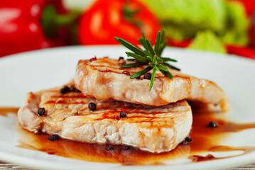 Pieces of meat grilled. Steak with rosemary and pepper in a white plate. Meat dish with red sauce on a white wooden table. Fresh tomatoes, peppers and cabbage near the plate.