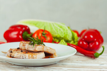 Pieces of meat grilled. Steak with rosemary and pepper in a white plate. Meat dish with red sauce on a white wooden table. Fresh tomatoes, peppers and cabbage near the plate.