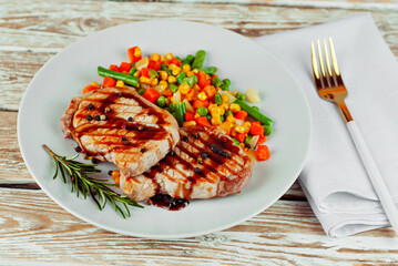 Pieces of meat grilled. Steak with rosemary and pepper in a white plate. Meat dish with red sauce on a white wooden table. Salad of corn, carrots and green peas near the steak.