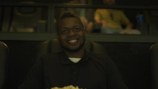 African American man sitting in armchair watching a comedy at the cinema, eating popcorn