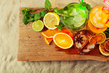 Orange and green cocktails on a wooden board. Alcoholic drink with ice, orange, mint and lime on the sand. A mixture of different drinks with fruit.