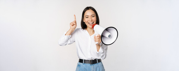 Smiling happy asian girl talking in megaphone and pointing up, announcing discount promo, showing...