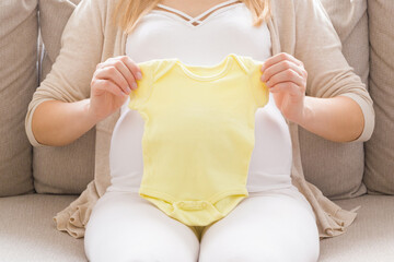 Young adult pregnant woman sitting on sofa at living room. Hands holding yellow bodysuit on big belly for future newborn. Preparing clothes in pregnancy time. Baby expectation. Front view. Closeup.
