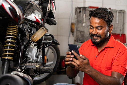 Young smiling mechanic using mobile phone in front of motors in showroom - concept of relaxation, taking break time and reading messages