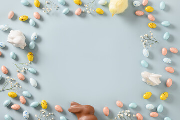 Easter border with pastel chocolate eggs, bunny, spring blossom flowers, and yellow chick on blue background. Top view. Flat lay. Happy Easter greeting card. Copy space.