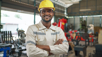Close up portrait of confident Africa American engineer man wearing safety helmet standing in the robotic arm manufacturing. Looking forward and crossed arm. Technology and industrial concept..