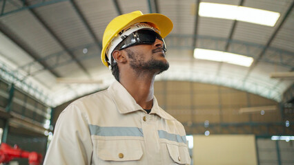 Close up portrait of confident Africa American engineer man wearing safety helmet and black glasses standing in the robotic arm manufacturing. Looking forward. Technology and industrial concept