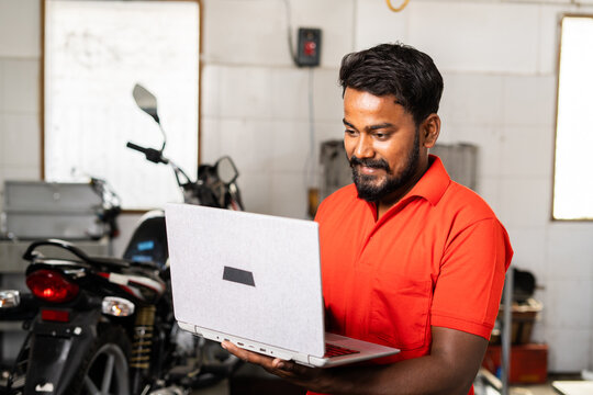Smiling motorbike mechanic busy using laptop at garage - concept of technology, checking online booking and maintenance and repair service.