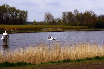 Pescador deportivo en un pequeño barco de pesca moderno. Pescador en uno de los muchos canales de Holanda rodeado de campos de cultivo. - obrazy, fototapety, plakaty