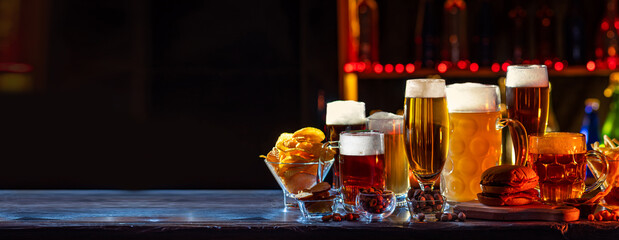 Glass of beer on bar counter. Jugs, mugs, pints of brew beverage, ale, cider on wooden table in pub, bar. Backlit dark showcase with craft beer bottles in brewery. French fries fried potatoes snack