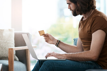 Time for some retail therapy. Shot of a handsome young man holding a credit card and using his laptop while relaxing in his living room.