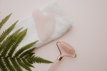 Gua sha and massage roller on a towel and fern on a pink background