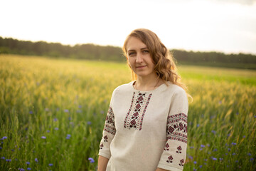 The girl is standing in the rye field. The young girl is in the green rye field. Woman is wearing Ukrainian national clothes, embroidered shirt. Smiling young lady with blue eyes, wavy blond hair.
