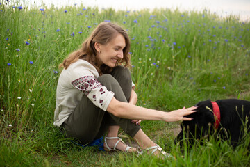 Fototapeta premium Girl wearing national Ukrainian clothes embroidered shirt is sitting in the beautiful rye field with her big black Labrador dog. 