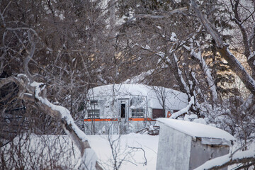 A small white with orange stripe retro metal camper surrounded by deep snow and forest of trees