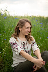 Girl wearing national Ukrainian clothes embroidered shirt is sitting in the beautiful rye field. Woman is holding blue field flowers cornflowers. Blue wildflowers and blond wavy haired girl
