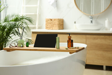 Wooden bath tray with tablet, face roller and cosmetic products on tub indoors