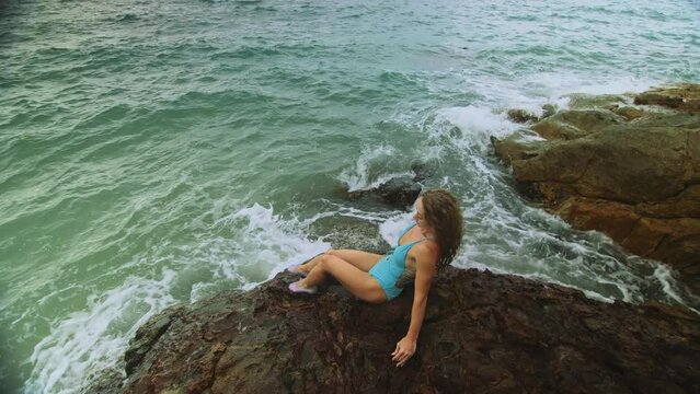 Alone attractive woman walk near a rock reef hill in stormy morning rain cloudy sea. Girl in turquoise swimsuit. Concept resort outdoor relax, vacation, loneliness. Dark dramatic view