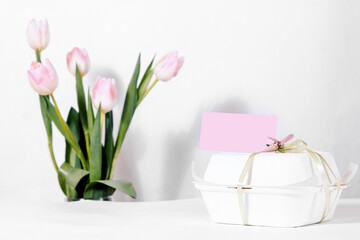 A small bento cake with a pink label in a box, next to tulips in defocus. White background for the eighth of March holiday. copyspace. Plastic lunch box with tasty bento cake and flowers