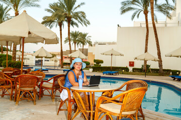 A young woman in a hat at a pool table at a hotel among palm trees drinks a cocktail and works remotely on a laptop.