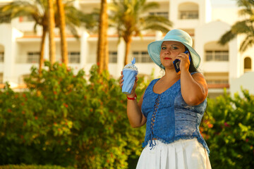 Naklejka premium A young woman in a blue hat among palm trees and greenery against the background of the hotel speaks on a mobile phone.
