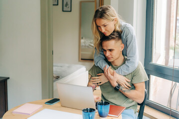 The girl hugs the guy while he works on the computer laptop at home.