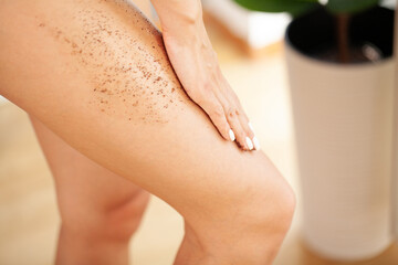 Beautiful young woman applying body scrub on leg at home - obrazy, fototapety, plakaty