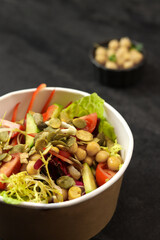 Vegetarian salad with chickpeas, tomatoes, cucumbers, bell peppers and pumpkin seeds in a round paper container on dark background.Healthy food, restaurant dish delivery. Top view. Free space for text