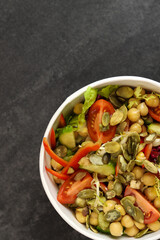 Vegetarian salad with chickpeas, tomatoes, cucumbers, bell peppers and pumpkin seeds in a round paper container on dark background.Healthy food, restaurant dish delivery. Top view. Free space for text