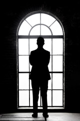 A silhouette of a man in a suit with his arms crossed facing an arched window with the light streaming in illustrating a decision, power and the unknown