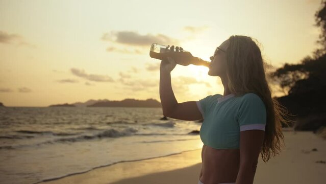 Sexy hot silhouette woman drinking whiskey with orange juice on beach. Rays gold sunset flare through glass. Woman in blue swimsuit, sunglasses. Concept happy summer holiday, alcohol drink, beverages