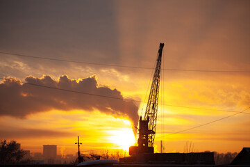 Obraz premium Against the background of an orange sunset, the outline of a gantry crane and smoke from a chimney of a heating plant.