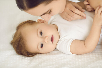 Little child together with mom, close up portrait. Happy beautiful young mother and her cute baby with soft fair hair cuddling on clean white sheets in comfy warm bed. Family, love, and care concept