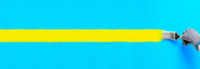 Hand holding paintbrush painting stripe yellow color on blue wall. Colorful art brush strokes...