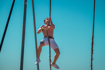 Strong, muscular, athletic young man climbing rope outdoor, working out in fresh air.