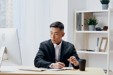 Fototapeta premium Asian man in a suit glass of coffee sits at a tablein front of a computer Workspace
