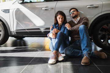 A sad young couple sits next to a car in a car dealership because they can't afford the car they want.