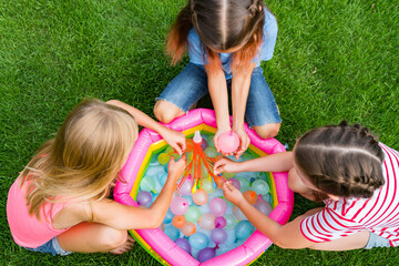Water balloon games for kids. Close up of girls filling up water balloons at sunny day. Summer fun outdoor activities for children concept - obrazy, fototapety, plakaty