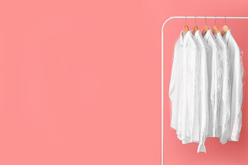 Rack with clean white shirts in plastic bags on pink background