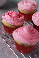 close up of vanilla muffins decorated with pink buttercream frosting on top in red baking cups, on grey marble table and white stone wall