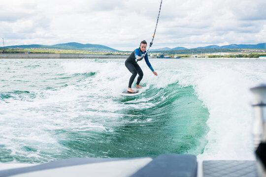 Female in wetsuit riding the waves using of tow rope behind a boat on sunny day. Watersport concept. Young athletic woman learning wakesurfing and perfecting tricks
