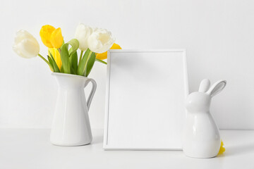 Blank photo frame, Easter rabbit and bouquet of tulips on table near white wall