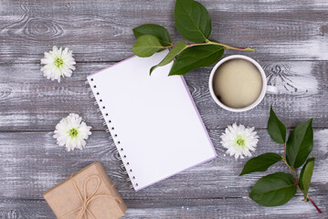 Top view of empty diary for ideas, plans, cup of coffee, gift wrapped in kraft paper, green branch and white chrysanthemum flowers on gray bleached wooden table. Copy space