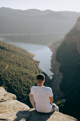 Vertical back view of a young man sitting on a cliff edge above a lake