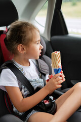 A child in a car seat, fastened with seat belts, holds a French bun with a sausage in his hands. The girl got hungry on the trip and eats a hot dog. Takeaway food. Food drive. 