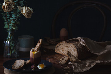 Breakfast table scene. Chiaroscuro style food photo. Cracked and open boiled egg with dippy bread set against a dark background with copy space available
