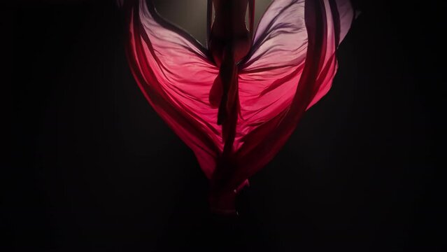 Young female circus gymnast spins on aerial silk and demonstrates grace of movement. Acrobat performs tricks at a height, red silk flutters like wings on black background with backlight. Slow motion.